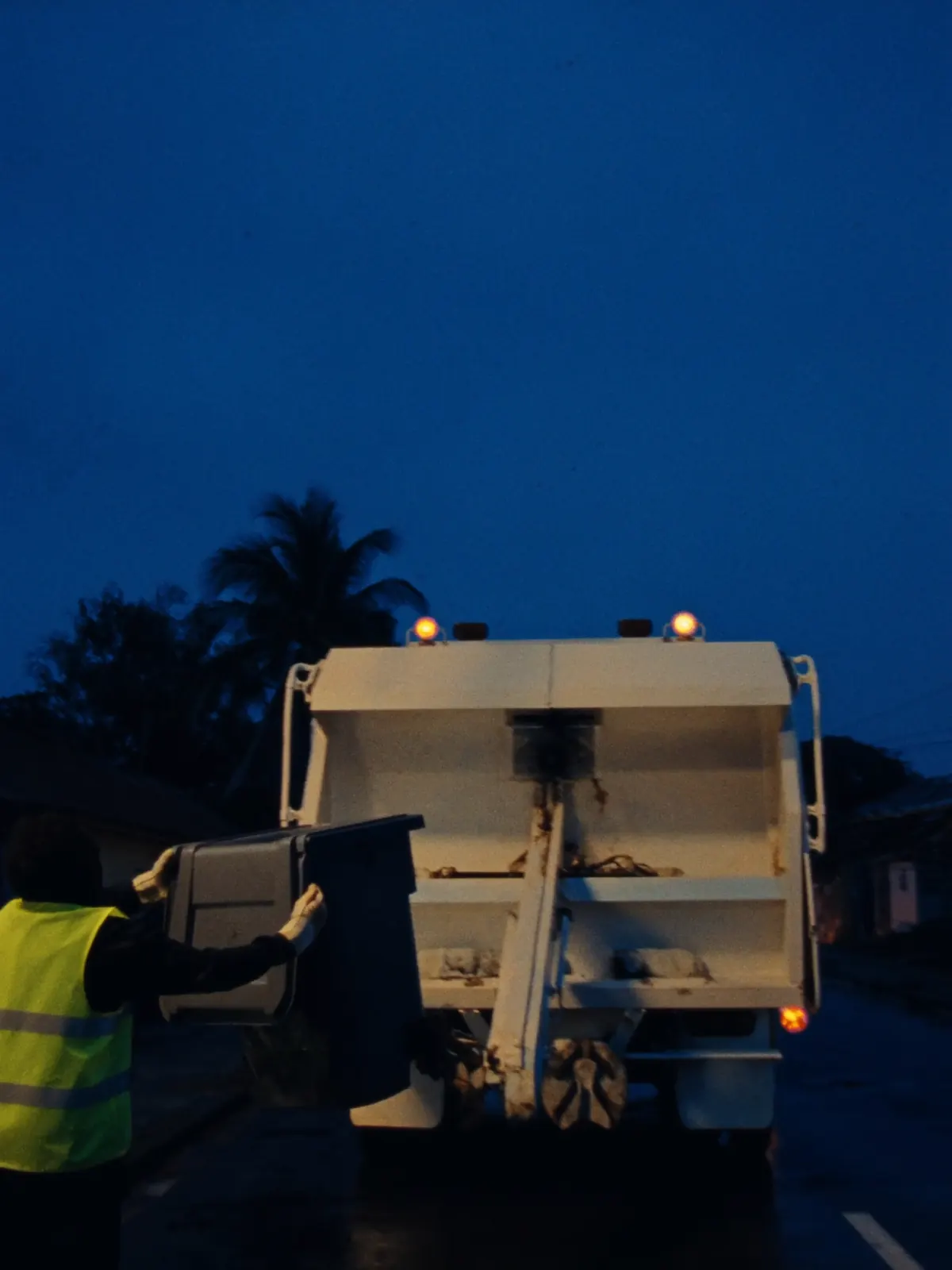 At dusk, a crew member tips a wheelie bin into the back of a refuse truck.