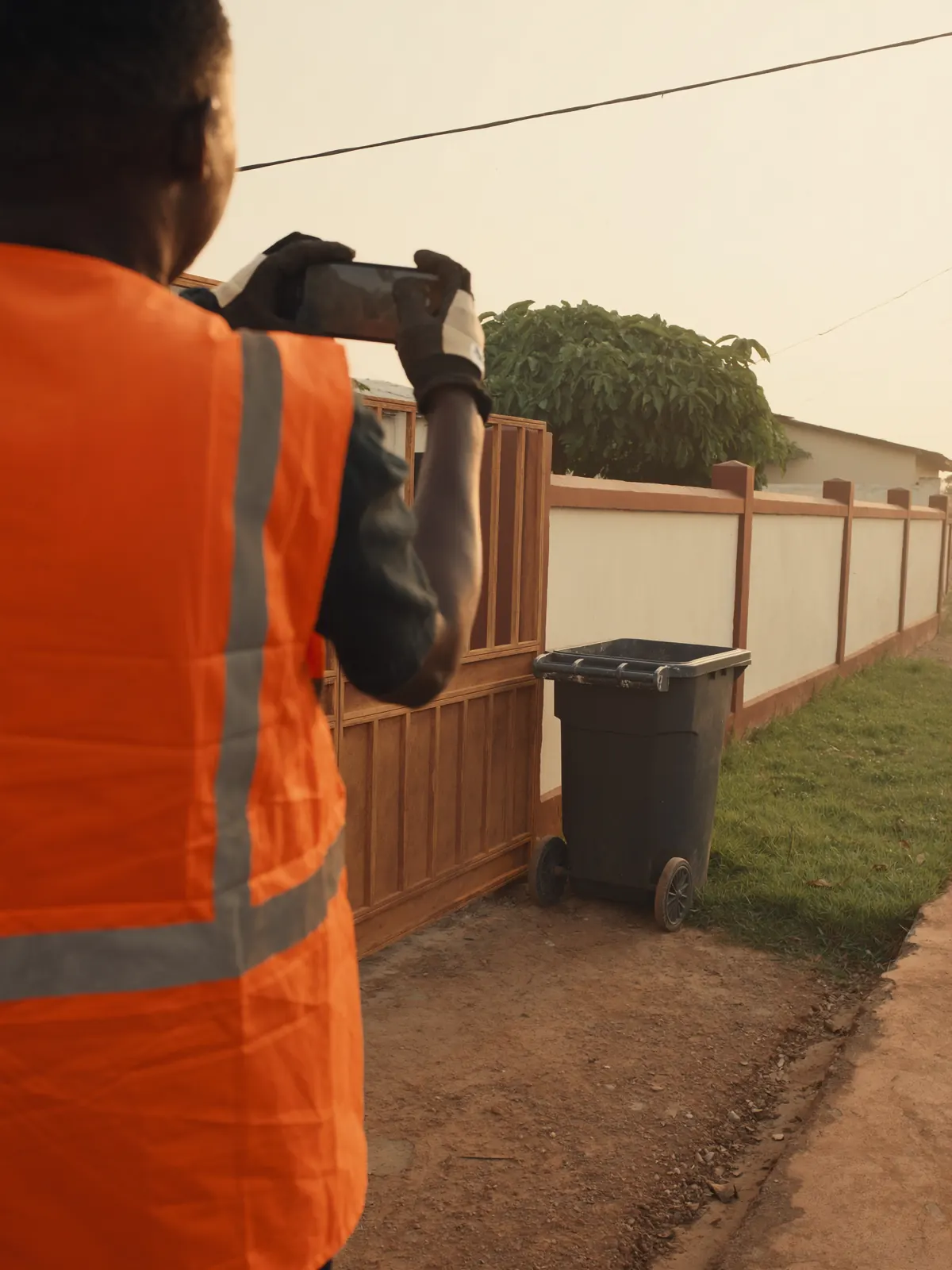 A crew member photographs an emptied wheelie bin at a compound gate.