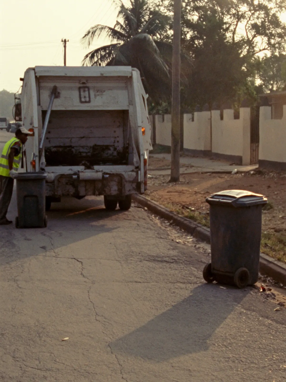 Wheelie bins wait outside homes on a street while a crew empties one into a truck in the distance.